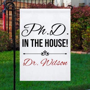 May include: White garden flag with black text that reads "Ph.D. in the house!" and "Dr. Wilson" in red text.