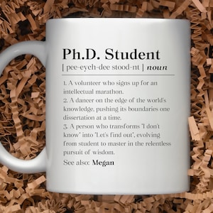 May include: White ceramic mug with a black and white design that defines a Ph.D. student as a volunteer who signs up for an intellectual marathon, a dancer on the edge of the world's knowledge, and a person who transforms "I don't know" into "Let's find out." The mug also says "See also: Megan".