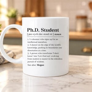 May include: A white ceramic mug featuring black printed text that defines a Ph.D. Student as a volunteer for an intellectual marathon, a dancer on the edge of knowledge, and a person who transforms I don't know into Let's find out. See also: Megan.