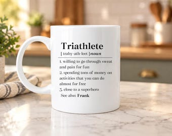 Puede incluir: Una taza de cerámica blanca con texto impreso en negro que define la palabra Triathlete como sustantivo, seguida de tres descripciones humorísticas numeradas y la frase See also: Frank, todo en una fuente sans-serif clara.