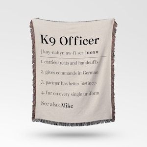 May include: A cream-colored woven throw blanket featuring black text that defines K9 Officer with a phonetic pronunciation, a noun label, and a numbered list of four humorous characteristics, finished with multicolored fringed edges on the sides.