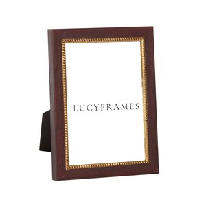May include: A dark brown picture frame with a gold trim and a beaded design. The frame is standing on a white surface. The text "LUCYFRAMES" is printed on the frame.