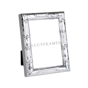 Puede incluir: Un marco de fotos plateado con un diseño de estilo bambú. El marco rectangular tiene un soporte negro en la parte trasera. El texto "LUCYFRAMES" está impreso en el centro del marco.