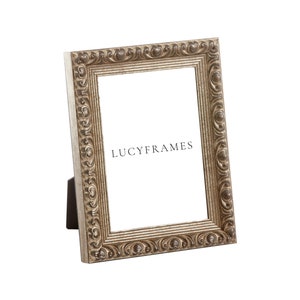 Puede incluir: Un marco de fotos plateado con un dise&ntilde;o ornamentado. El marco est&aacute; de pie sobre una superficie blanca. El texto "LUCYFRAMES" es visible en el marco.