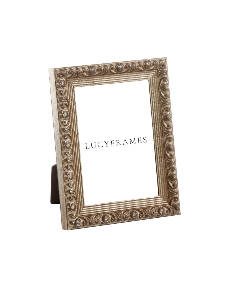 Puede incluir: Un marco de fotos plateado con un dise&ntilde;o ornamentado. El marco est&aacute; de pie sobre una superficie blanca. El texto "LUCYFRAMES" es visible en el marco.