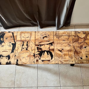 May include: Wooden coffee table featuring engraved portraits of characters from the anime series "One Piece." The table is made of light-colored wood and has black legs. The artwork is detailed and covers the entire surface.