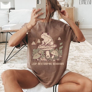 May include: A brown t-shirt with a graphic of a frog sitting on a mushroom. The text "Leap into shaping behaviors" is printed below the graphic.