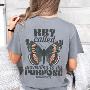 May include: Gray t-shirt with a butterfly graphic and the text "RBT called according to His PURPOSE Romans 8:28." The butterfly has green, orange, and black details. The shirt is a casual style.