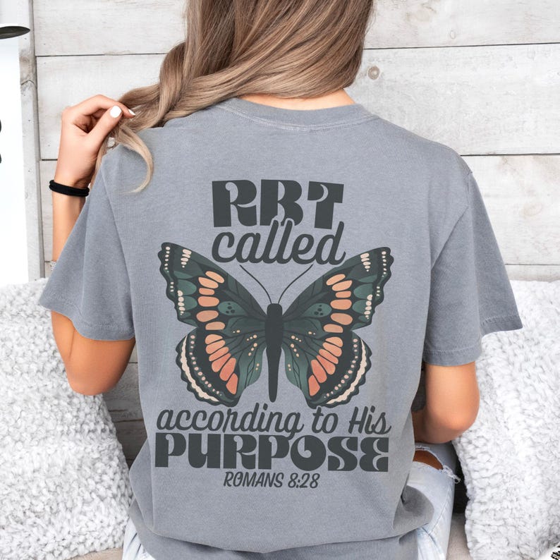 May include: Gray t-shirt with a butterfly graphic and the text "RBT called according to His PURPOSE Romans 8:28." The butterfly has green, orange, and black details. The shirt is a casual style.
