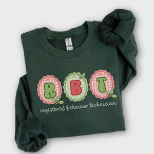May include: A dark green crewneck sweatshirt featuring the letters R, B, and T in a bubble font, each centered within a scalloped gingham circle. The text "registered behavior technician" is printed below in white, accented with holly leaves.