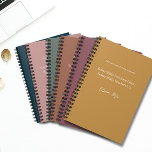 May include: A set of six spiral bound journals with different colored covers. The covers are teal, pink, green, burgundy, and brown. The front cover of the brown journal has the text "The Shift Reflection Journal" and the text "Some Shifts You Save Lives, Some Shifts You Survive." with the name "Olivia Rh" written below.