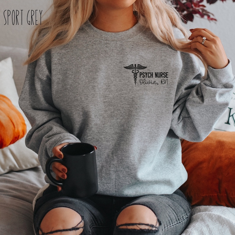 May include: A gray sweatshirt with a black graphic of a caduceus and the text "PSYCH NURSE Olivia, RN".