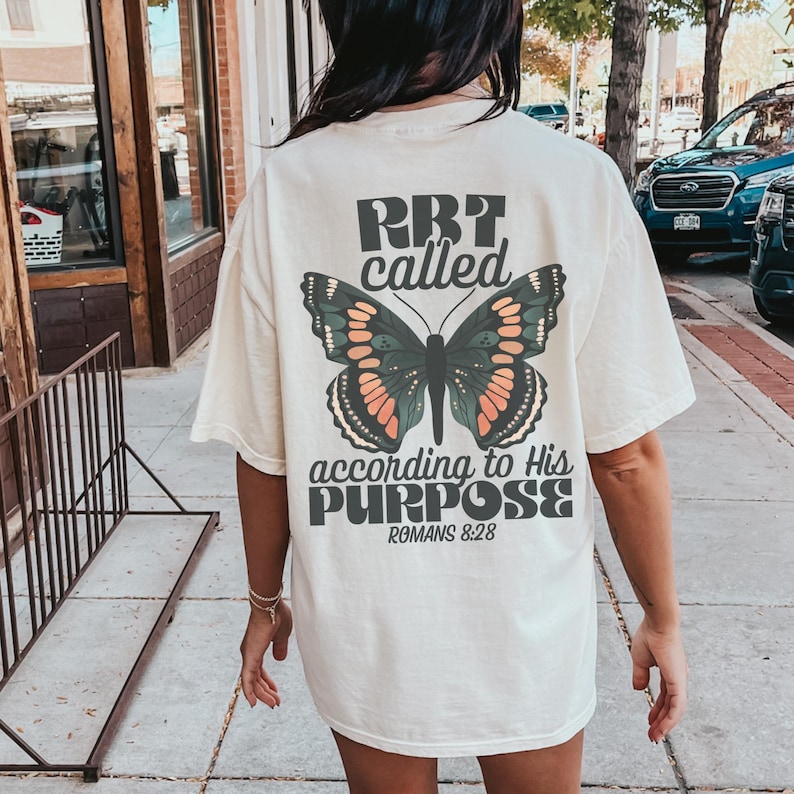 May include: Cream-colored t-shirt with a large butterfly graphic and the text "RBT called according to His PURPOSE Romans 8:28." The butterfly has orange, green, and black details. The shirt is a casual, oversized style.