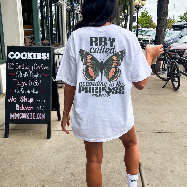 May include: White t-shirt with a butterfly graphic and the text "RB7 called according to His PURPOSE ROMANS 8:28". The shirt is worn outdoors. A sign for cookies is visible in the background.