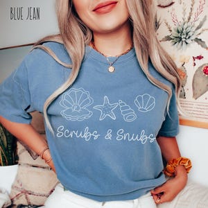 May include: Blue jean colored t-shirt with white seashell graphics and the words "Scrubs & Snubs". The top has short sleeves and a crew neck. The text "BLUE JEAN" is in the upper left corner.