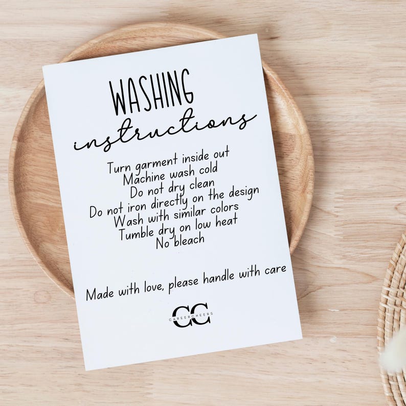 May include: A white card with black text, featuring washing instructions. The card is on a wooden plate. Text includes "Turn garment inside out", "Machine wash cold", and "Made with love, please handle with care."