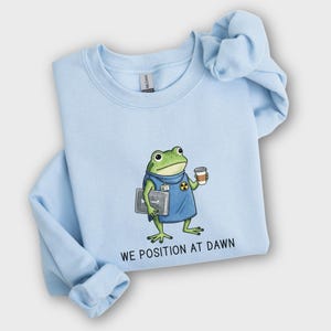 May include: A light blue crewneck sweatshirt featuring a graphic of a green frog wearing a blue medical scrub top with a radiation symbol, holding an X-ray film and a coffee cup, with the text "WE POSITION AT DAWN" printed in black below the frog.