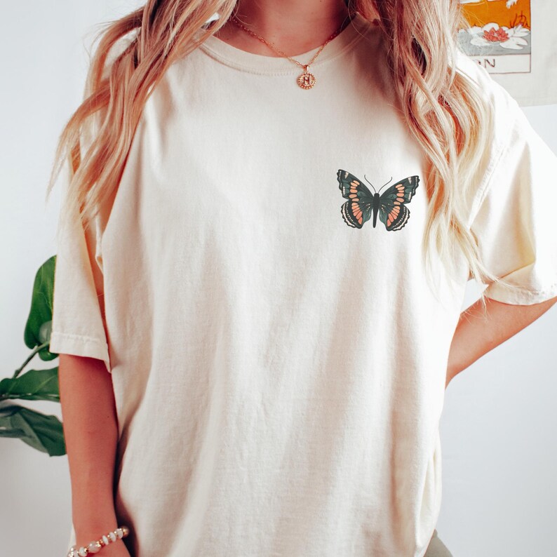 May include: Cream-colored t-shirt featuring a butterfly graphic. The butterfly design is in shades of black, coral, and green. The shirt has short sleeves and a relaxed fit, suitable for casual wear.