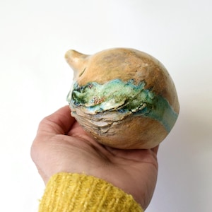 May include: A small, round, ceramic vase with a textured surface. The vase is decorated with a band of blue and green glaze.
