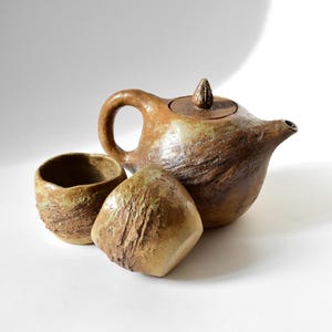 Può includere: Una teiera in ceramica marrone e beige con un coperchio e due tazze abbinate. La teiera e le tazze hanno una finitura rustica e testurizzata.