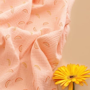 May include: A pink fabric with a repeating pattern of watermelon slices. The fabric is soft and textured, and the watermelon slices are a light orange and yellow colour.
