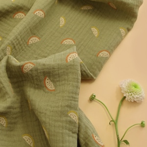 May include: A green fabric with a repeating pattern of orange and yellow watermelon slices. The fabric is folded and draped over a light brown surface.