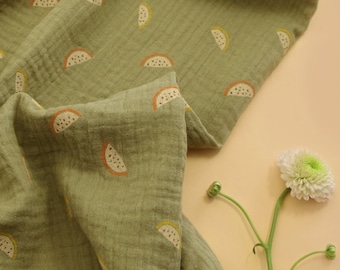 Pode incluir: Um tecido verde com um padrão repetido de fatias de melancia laranja e amarela. O tecido está dobrado e drapeado sobre uma superfície marrom clara.