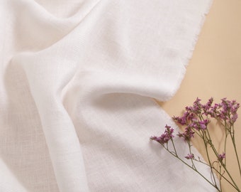 Könnte beinhalten: Weißer Stoff mit einer weichen, strukturierten Oberfläche. Der Stoff ist drapiert und gefaltet, wodurch ein glatter, fließender Look entsteht. Ein paar kleine, rosafarbene Blütenzweige liegen auf dem Stoff.