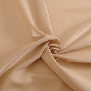 May include: A close-up of a smooth, light beige fabric. The fabric is folded and draped, showcasing its soft texture and subtle sheen.