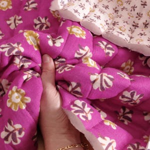 Pode incluir: Close-up de um tecido acolchoado com um padrão floral. O tecido é de uma cor fúcsia vibrante com desenhos de flores brancas, amarelas e roxas. O acolchoado cria um efeito fofo e texturizado. Uma porção de um tecido floral branco é visível.