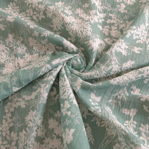 Könnte beinhalten: Nahaufnahme eines Stoffes mit floralem Muster. Der Stoff ist ein weiches, gedämpftes Grün mit einem weißen Blumendesign. Der Stoff ist in einer Spirale zusammengefasst, wodurch die Textur und das Muster des Materials sichtbar werden.