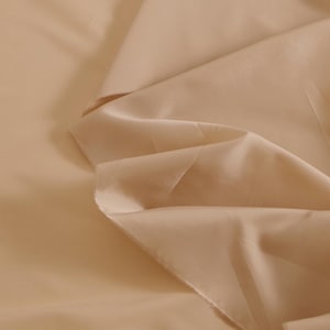 May include: A close-up of a smooth, light beige fabric with a soft, flowing texture. The fabric is folded and draped, showcasing its delicate drape and subtle sheen.