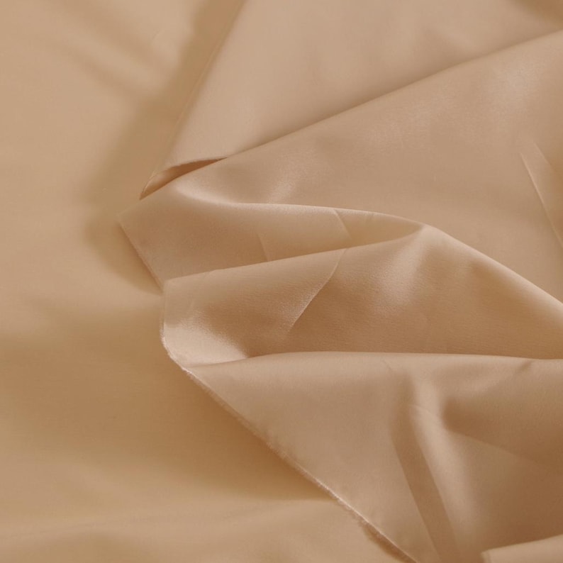 May include: A close-up of a smooth, light beige fabric with a soft, flowing texture. The fabric is folded and draped, showcasing its delicate drape and subtle sheen.