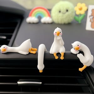 Peut inclure: Ensemble de décorations blanches en forme d'oie pour les bouches d'aération de voiture, avec des becs et des pattes oranges. Les oies sont dans diverses poses, dont allongées, assises et pliées. Autres décorations : arc-en-ciel, boule verte et fleur jaune.