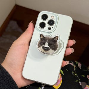 May include: A light green phone case with a cat face pop socket. The cat has black and white fur and is smiling. The phone case has a camera and flash. The phone is being held by a person.