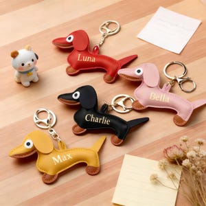 May include: A collection of leather dachshund keychains in red, pink, black, and yellow. Each keychain features a gold keyring and a personalized name. A small white and brown figurine is also present.