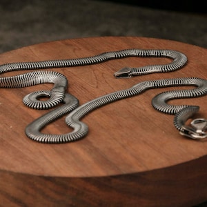 May include: Two silver-coloured snake necklaces rest on a round, wooden surface. The snakes have a ribbed texture, and one has a detailed snake head. The wood has a rich, warm tone, contrasting with the cool metal.