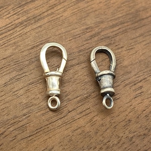 Könnte beinhalten: Zwei silberfarbene Metallverschlüsse mit einer Schlaufe unten. Die Verschlüsse befinden sich auf einer braunen Holzoberfläche.