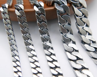 May include: Close-up of several silver-colored curb chains of varying widths. The chains are arranged diagonally, showcasing their interlocking links. The background is a light surface with a wooden bowl.