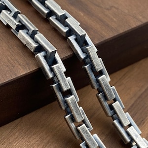 May include: Two silver chains with a box link design. The chains are laying on a wooden surface.