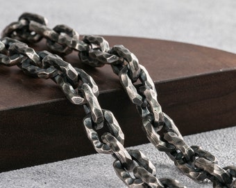 May include: A silver chain necklace with a chunky, textured link design. The chain is lying on a dark brown wooden surface.