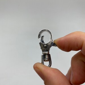 May include: A silver-colored swivel clasp with intricate engraved details. The clasp features a spring-loaded hook and a rotating base. The design includes dotted patterns and floral-like engravings, creating a decorative and functional accessory.