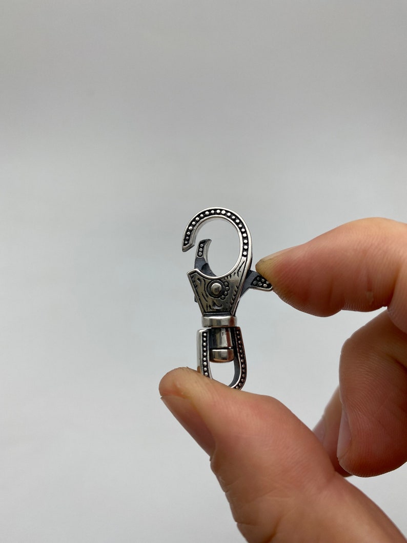 May include: A silver-colored swivel clasp with intricate engraved details. The clasp features a spring-loaded hook and a rotating base. The design includes dotted patterns and floral-like engravings, creating a decorative and functional accessory.