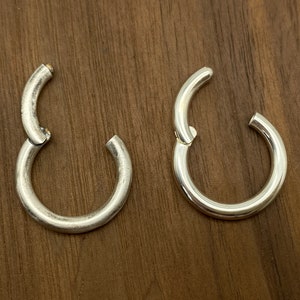 Könnte beinhalten: Zwei silberne Creolen, eine offen und eine geschlossen, auf einer Holzoberfläche. Die Ohrringe haben ein schlichtes, kreisförmiges Design mit einem Klappverschluss. Das Metall hat eine leicht gealterte, matte Oberfläche.