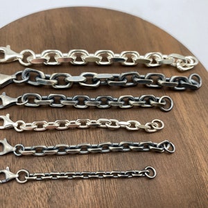 Könnte beinhalten: Ein Set aus sechs silbernen Kettenarmbändern in verschiedenen Längen und Gliedgrößen. Die Ketten liegen auf einer Holzoberfläche.