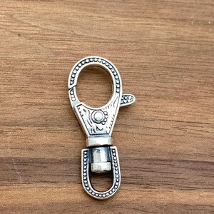 May include: A silver-colored swivel clasp with a decorative design. The clasp has a circular opening and a lever for easy attachment. The metal clasp is detailed with small circular accents and engraved patterns.