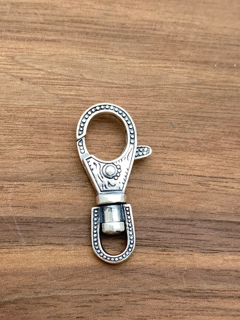 May include: A silver-colored swivel clasp with a decorative design. The clasp has a circular opening and a lever for easy attachment. The metal clasp is detailed with small circular accents and engraved patterns.
