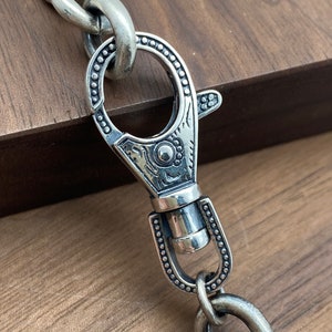 May include: Close-up of a silver-toned chain with a decorative clasp. The clasp features an ornate design with a circular opening and a spring-loaded lever. The chain links are large and oval-shaped, and the metal has a slightly aged appearance.