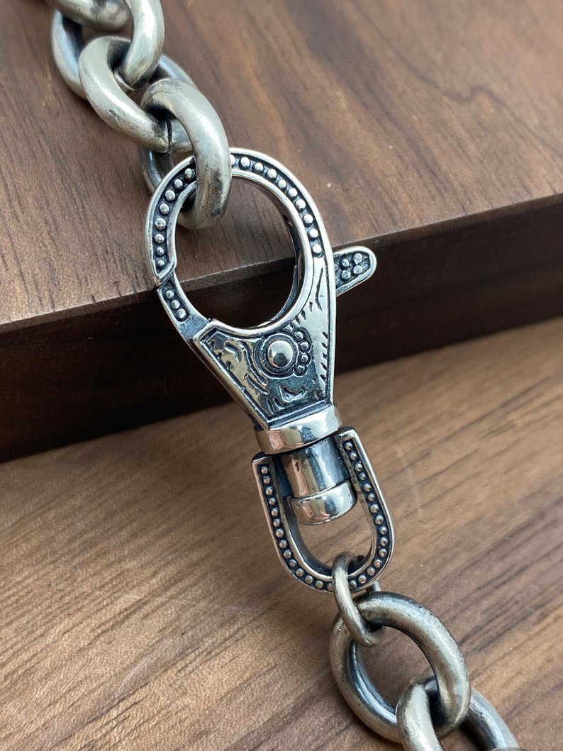 May include: Close-up of a silver-toned chain with a decorative clasp. The clasp features an ornate design with a circular opening and a spring-loaded lever. The chain links are large and oval-shaped, and the metal has a slightly aged appearance.