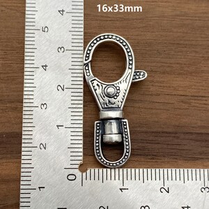 May include: A silver-colored swivel clasp with an ornate design, measuring 16x33mm. The clasp features a decorative pattern around the edges and a spring-loaded lever for secure closure. The image includes a ruler for scale.